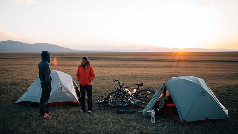 尼科尔和一个同伴站在草地上的白色小帐篷和一堆自行车中间, 太阳从远处冉冉升起
